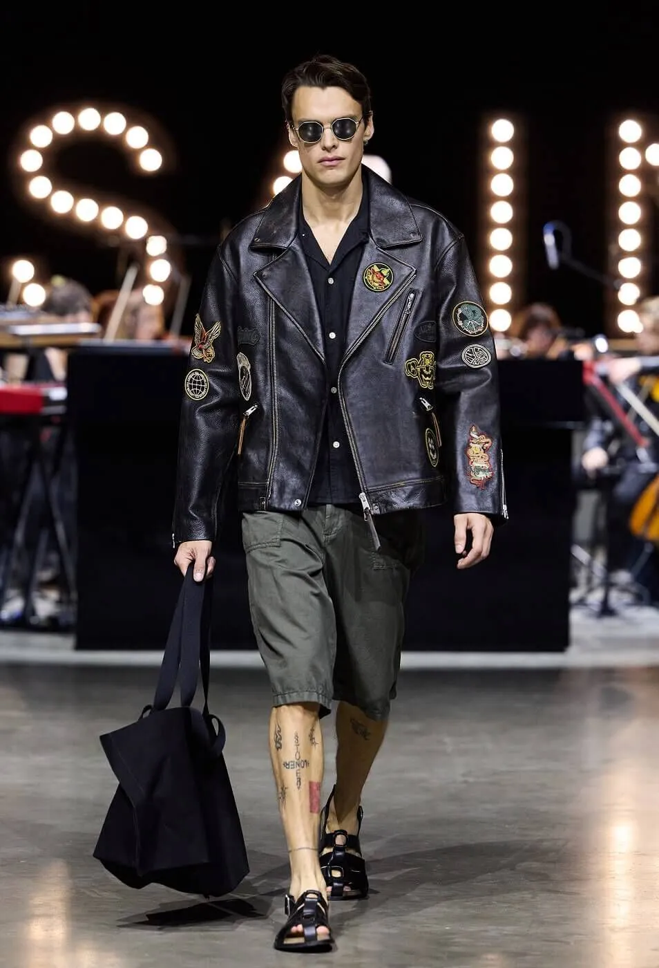 Man wearing a black leather jacket with patches,sunglasses and grey shorts