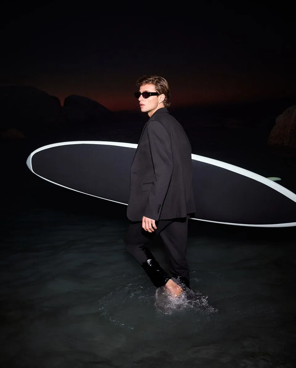 Photograph of a male model in a suit and holding a surfboard on the beach.