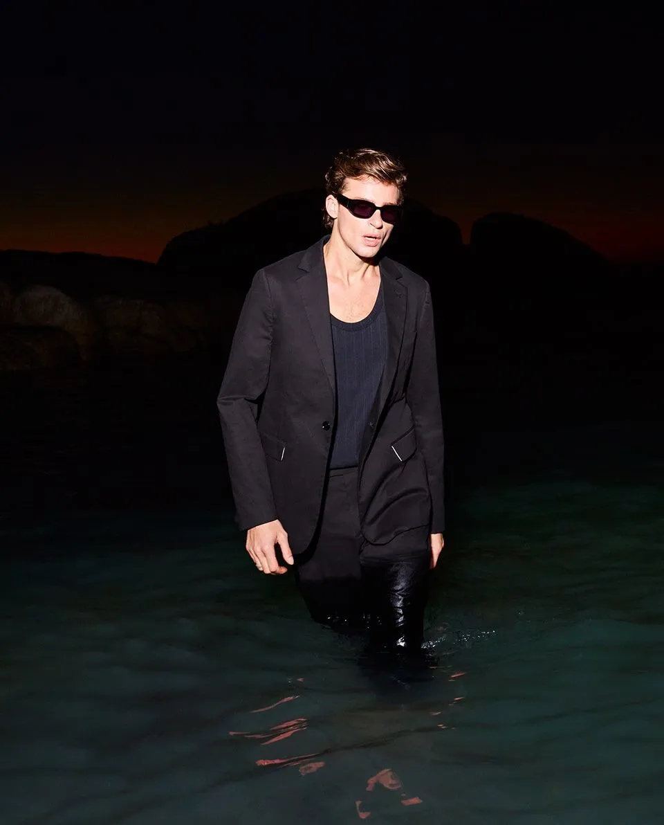 Photograph of a male model in a suit and holding a surfboard on the beach.