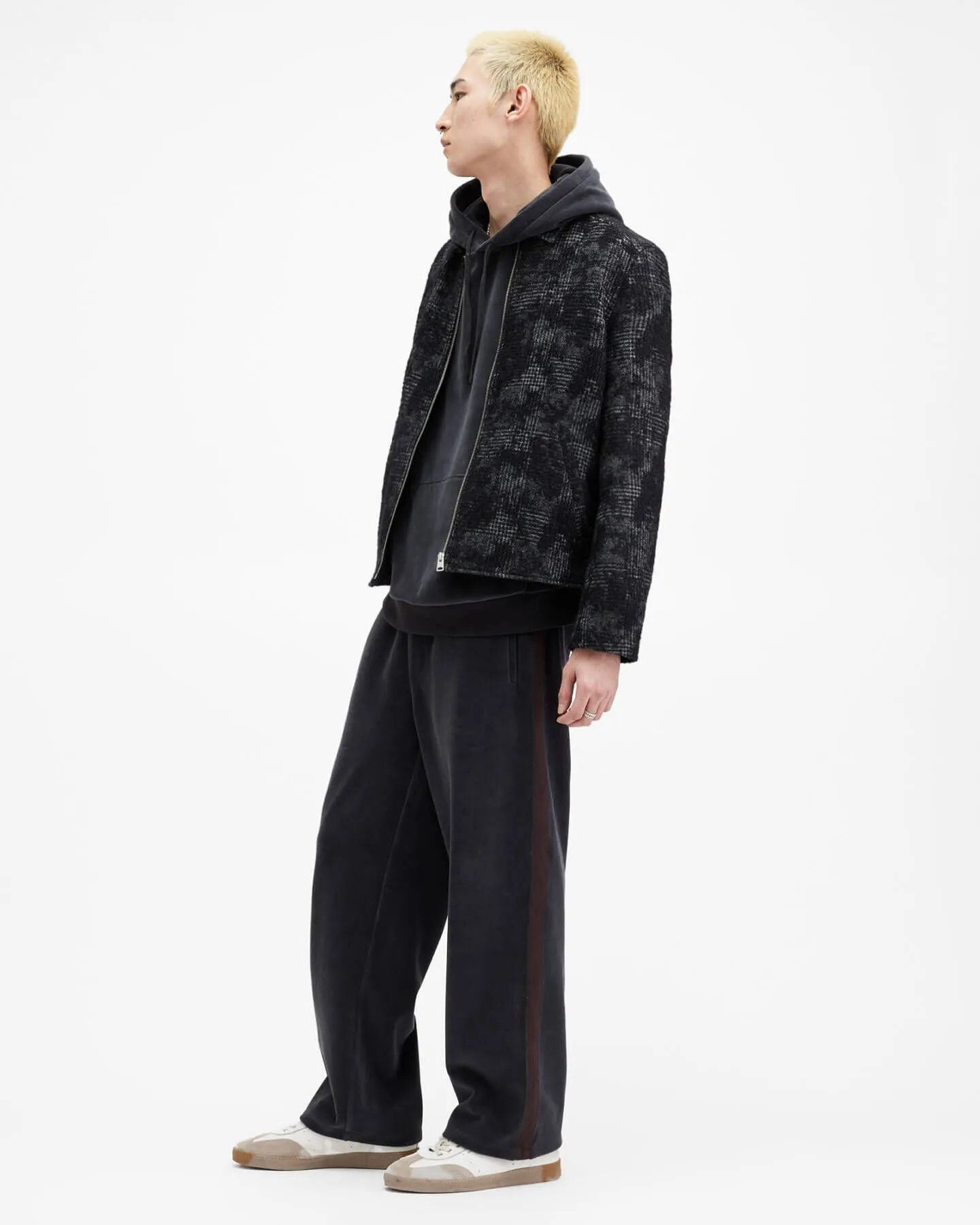 A man wearing a patterned jacket over a dark grey hoodie, with grey tracksuit bottoms and trainers.