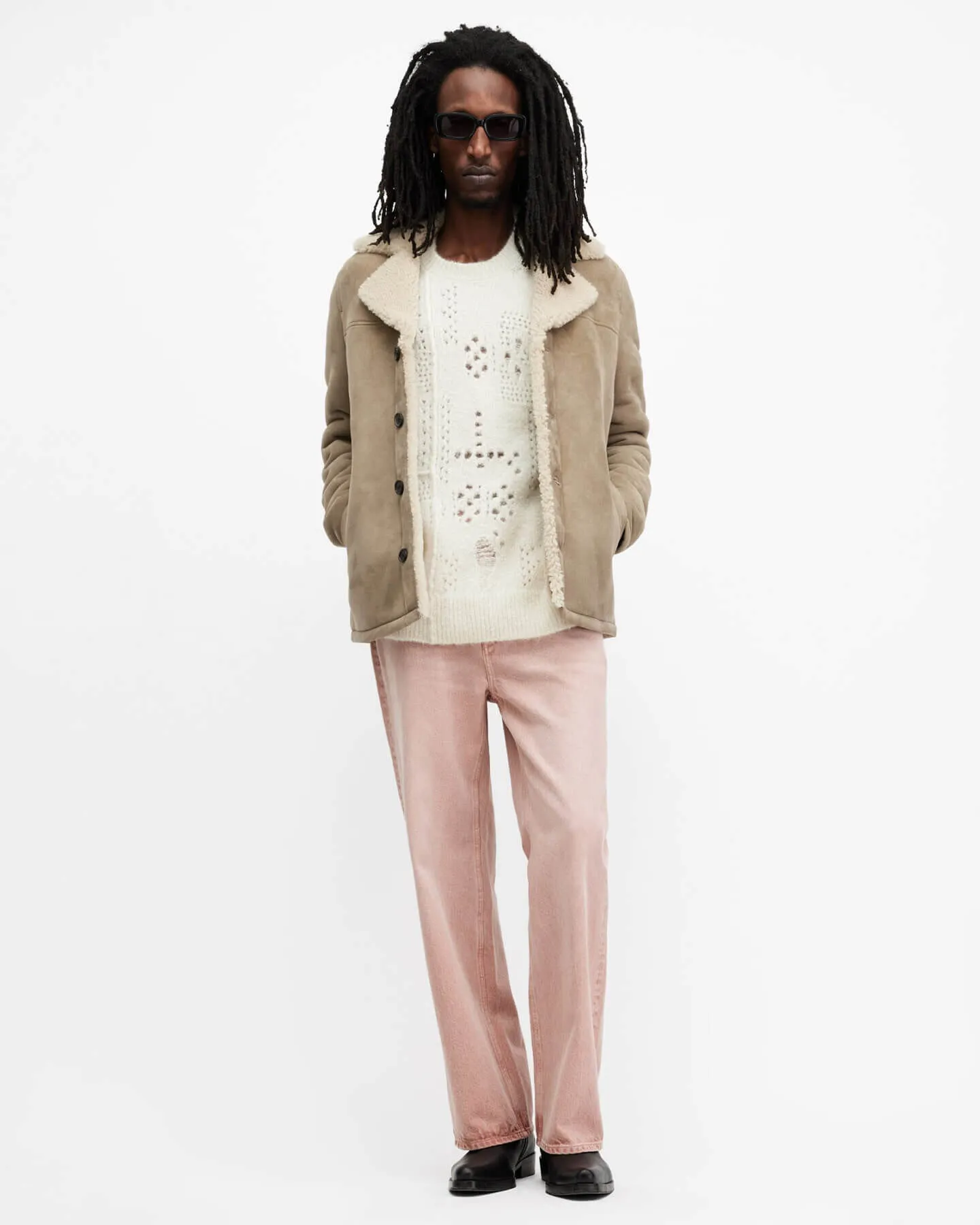 Man wearing a light brown shearling jacket over a holey cream jumper, with light pink jeans.